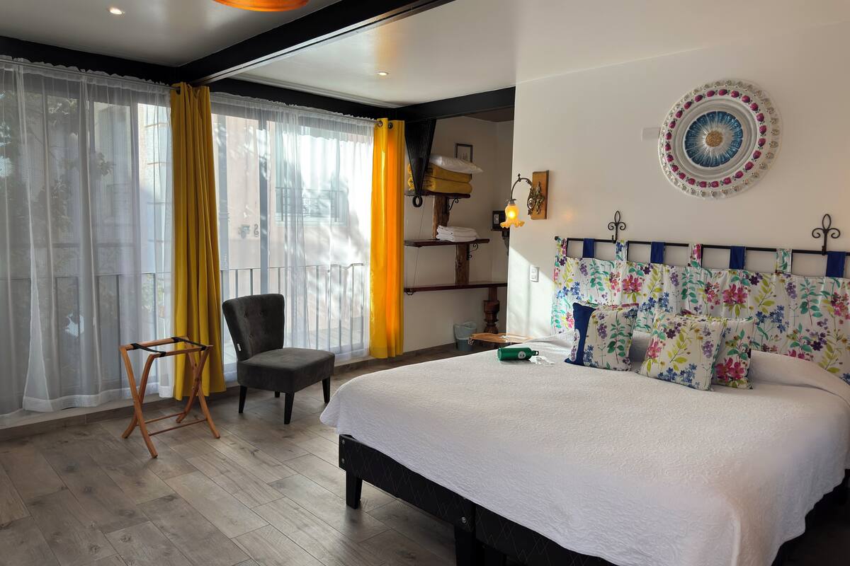 A bedroom with a bed, a chair, a window with curtains, a wall-mounted lamp, and a decorative plate.