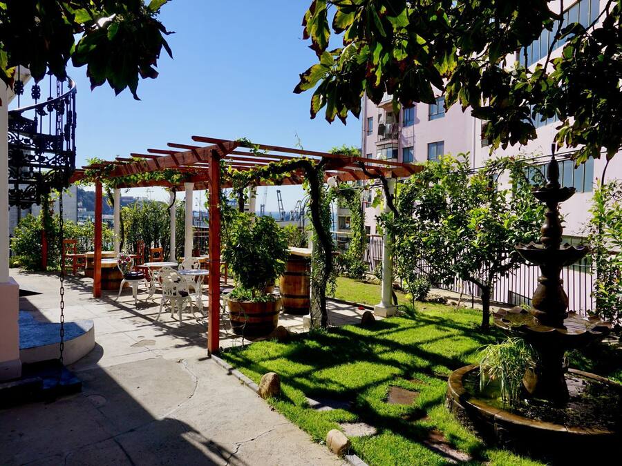 Un jardín con pérgola, plantas en macetas, una fuente y mobiliario exterior.