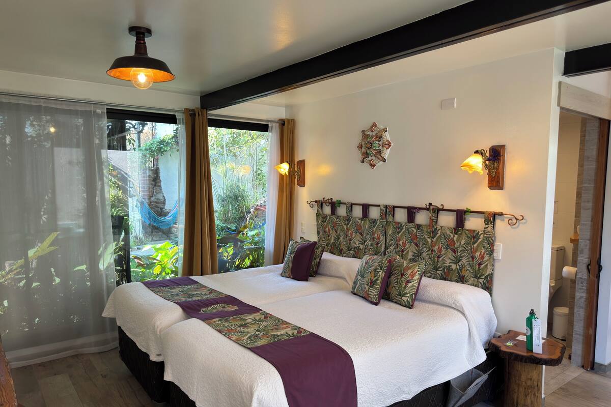 A large bed with a floral headboard, a bedside table with a green bottle, and a view of greenery outside.