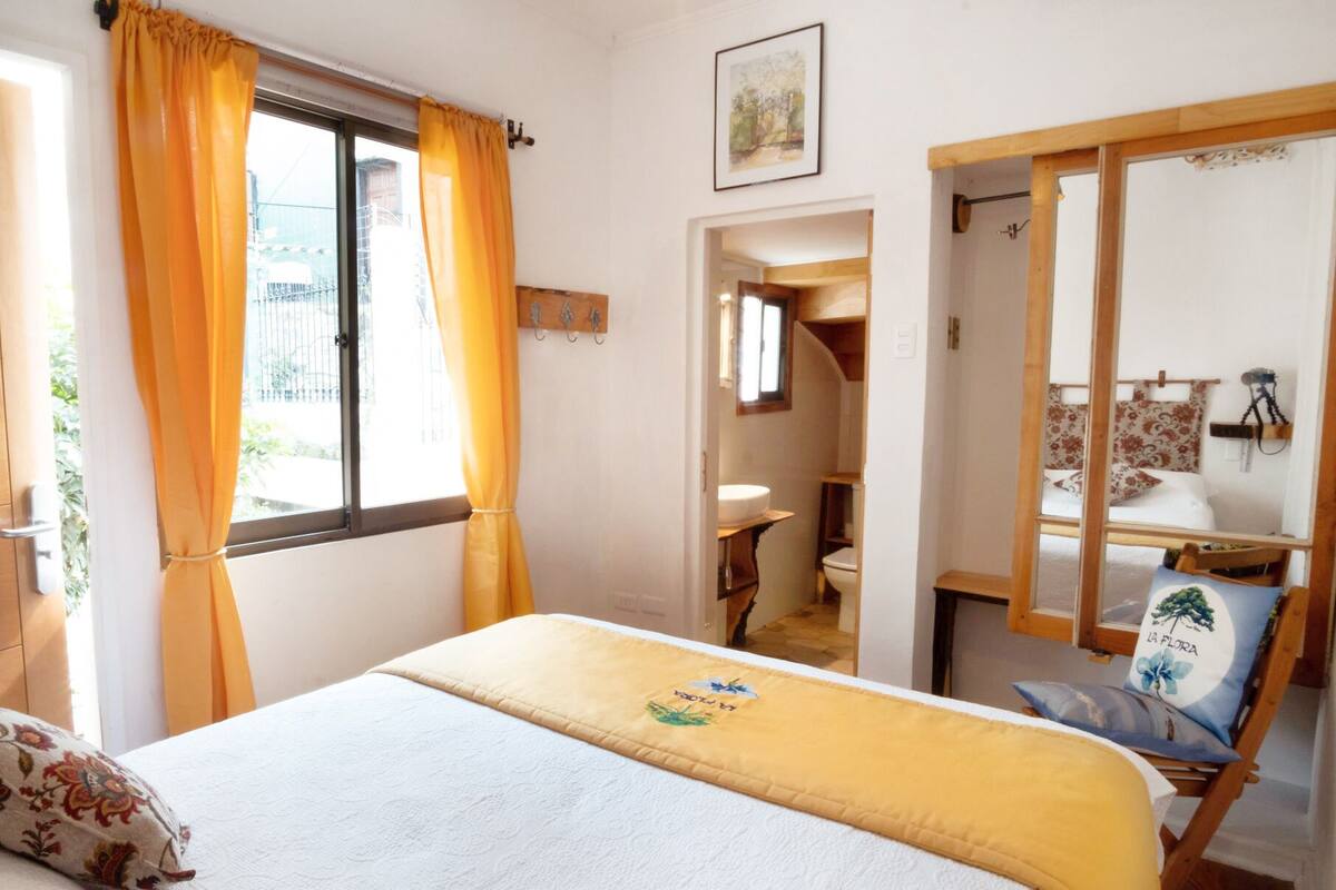 A bedroom with a bed, a window with curtains, a bathroom visible through an open door, and a small seating area with a chair and cushions.