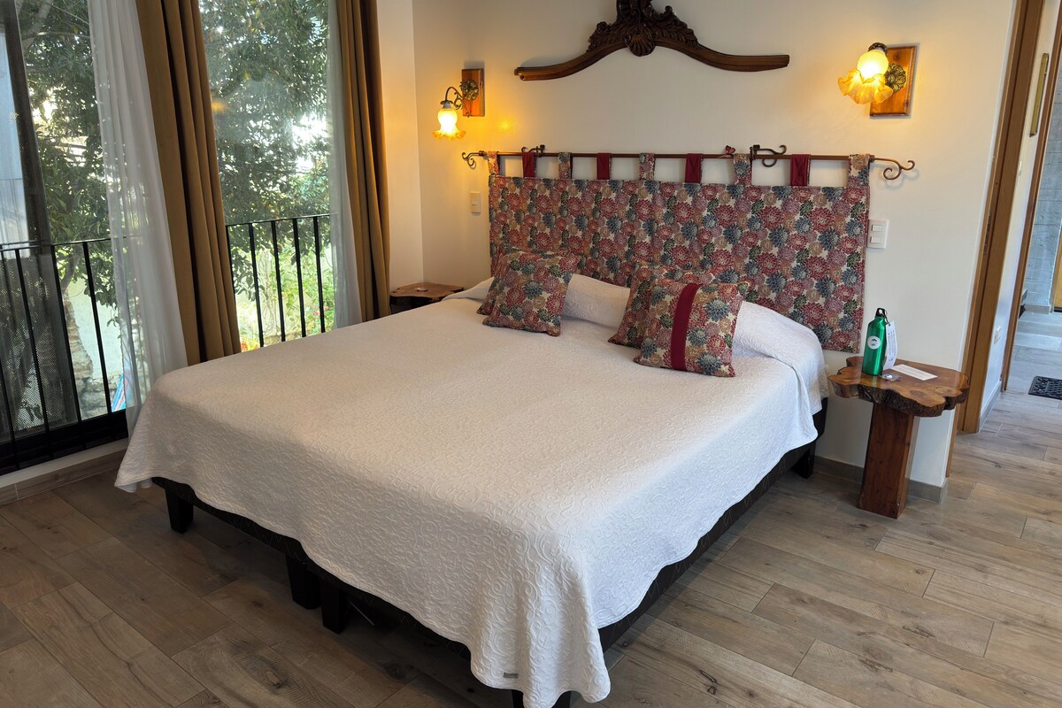 Een ruime slaapkamer met een groot bed, een hoofdeinde met bloemenmotief, twee bedlampen en uitzicht op groen via een schuifdeur.