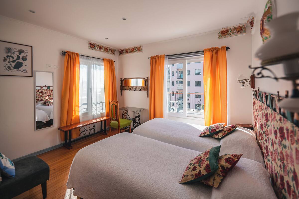 A hotel room with two beds, a bench, a small table, and a window with curtains.