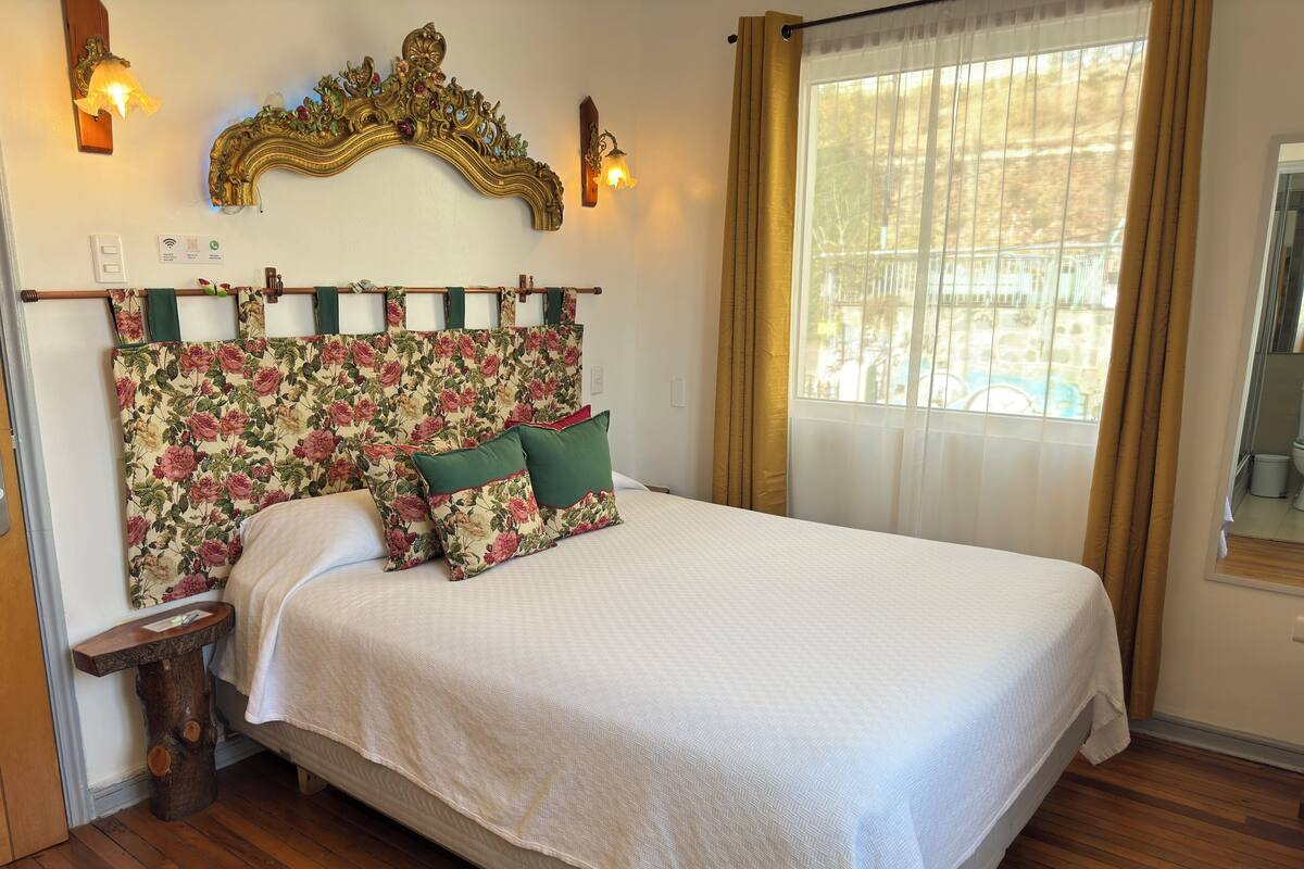 A bed with a floral headboard, a wooden nightstand, and a window with curtains.