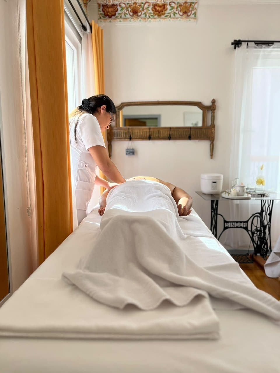 Ein Massageräum mit Liege, Massagetisch, Spiegel und einem kleinen Tisch mit Tasse und Untertasse.