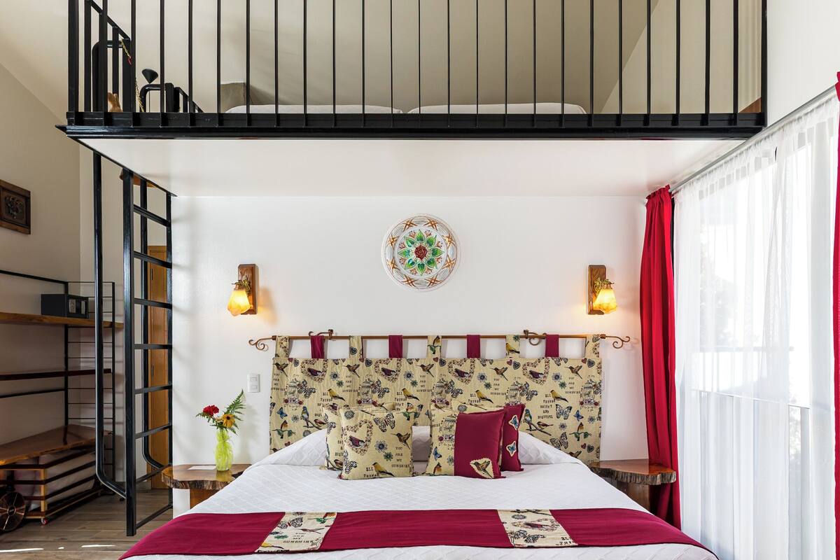 A bedroom with a bed, a headboard with a decorative pattern, a window with curtains, a staircase, and a wall-mounted light fixture.