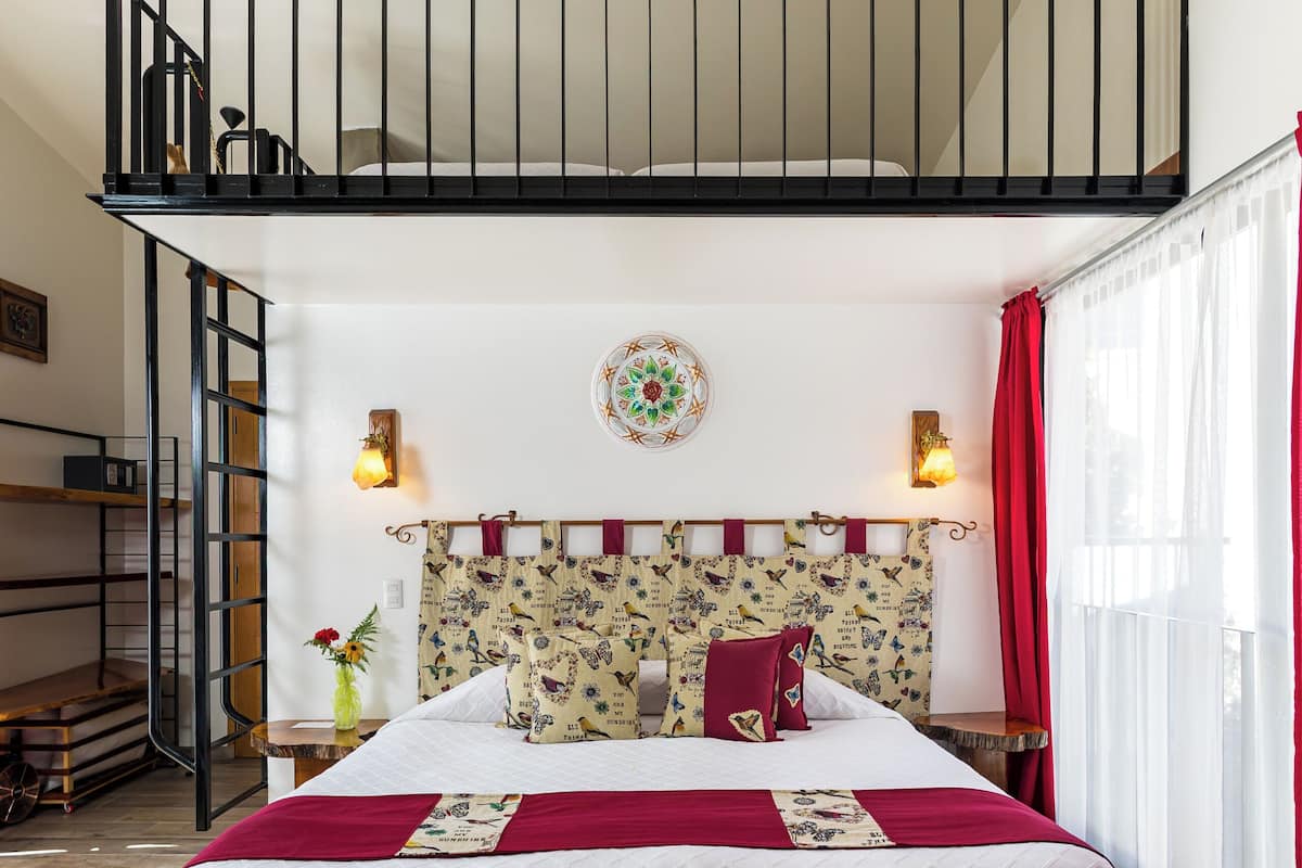 A bedroom with a bed, a headboard with a decorative pattern, a window with curtains, a staircase, and a wall-mounted light fixture.