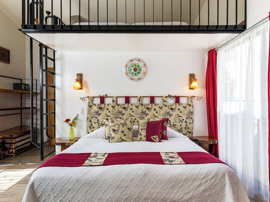 Un dormitori amb un llit, un capçal amb un dibuix decoratiu, una finestra amb cortines, una escala i una làmpada penjant a la paret.