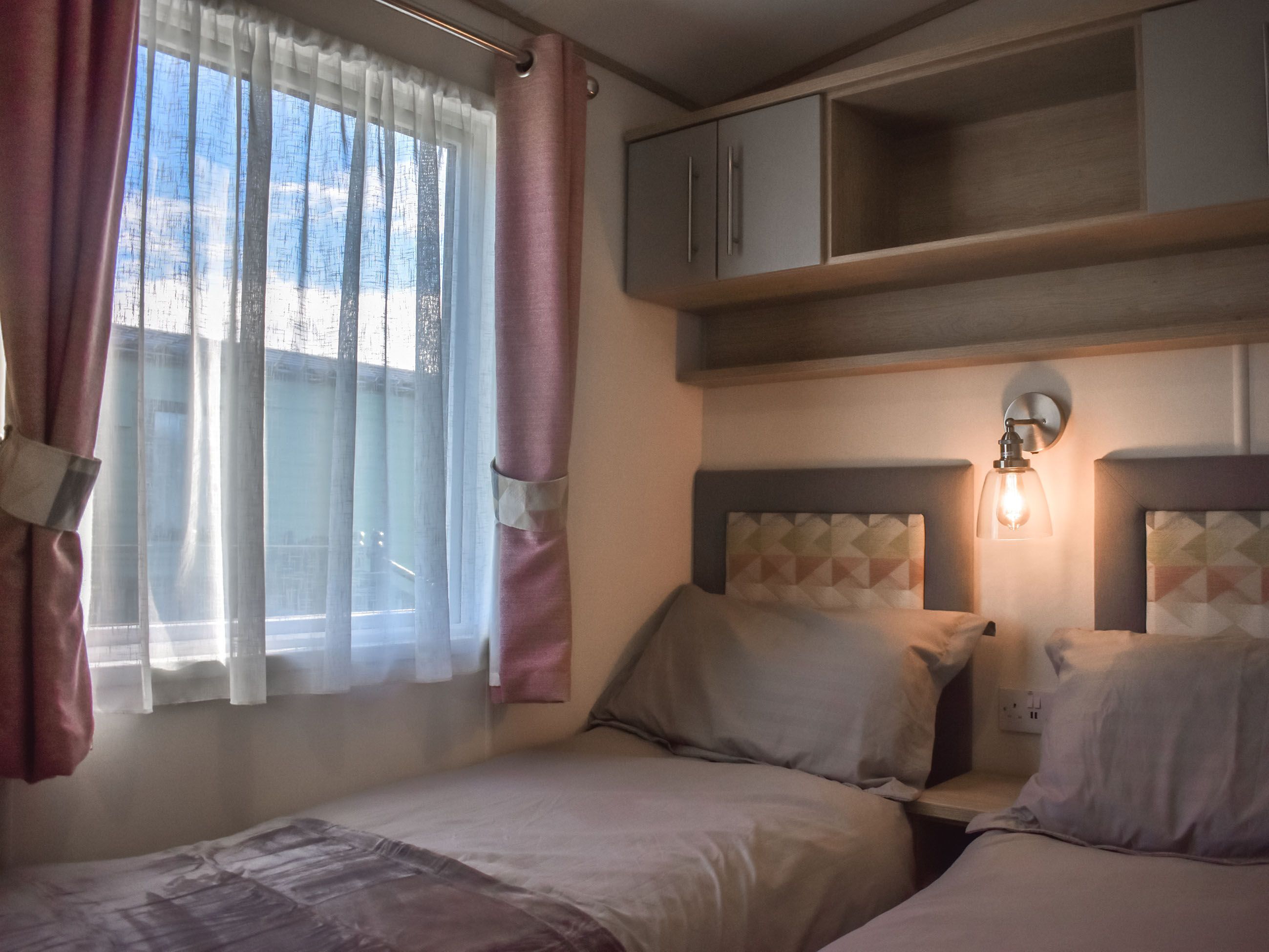 A bedroom with two beds, a window with sheer curtains, a headboard with a light fixture, and a wooden shelf above the beds.