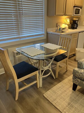 A dining area with a glass table, two chairs, a kitchen in the background, and a window with blinds.