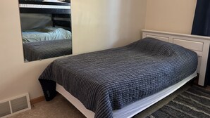 A single bed with a dark patterned bedspread, a white headboard, and a wall-mounted mirror reflecting the bed.