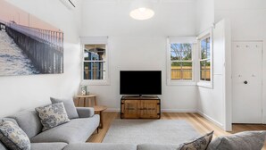 Uma sala de estar com um sofá cinzento, uma TV de tela plana sobre um móvel de madeira, uma mesa pequena com um jarro e um quadro de uma ponte.