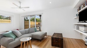 A living room with a grey sectional sofa, a small round table, a wooden chest, a ceiling fan, and a TV on a white shelf.