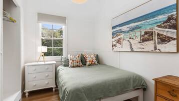 Un dormitorio con cama, mesita de noche, lámpara, ventana y una pintura de una escena de playa.