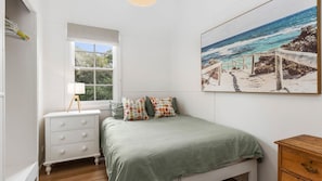 A bedroom with a bed, a bedside table, a lamp, a window, and a beach scene painting.