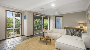 A modern living room with a beige sofa, a coffee table, and a view of lush greenery through large sliding glass doors.