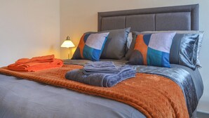Een netjes opgemaakt bed met een grijze hoofdbord, meerdere kussens in verschillende kleuren en een opgevouwen oranje handdoek op het bed.