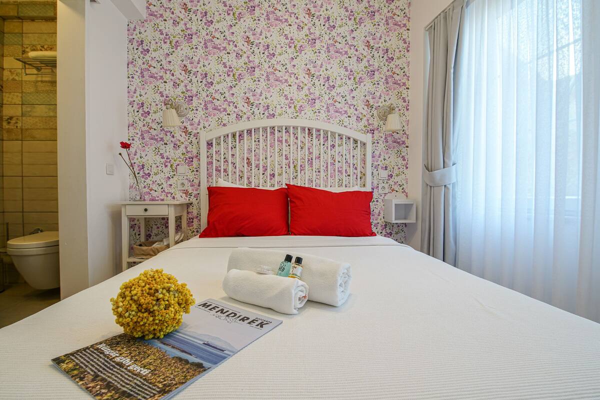 A neatly made bed with red pillows, a floral wallpaper, and a small bedside table with a vase of flowers.