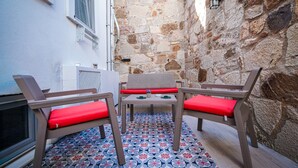 Uma pequena área de estar ao ar livre com duas cadeiras e uma mesa, sobre um piso estampado, em frente a uma parede de pedra e um edifício branco.