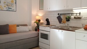 Ett kompakt kök med vita skåp, en spis och ett handfat. Det finns en liten matplats med ett bord och en soffa.