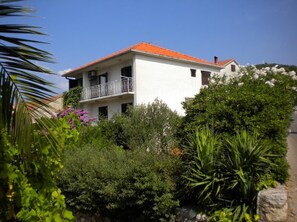 A two-story building with a red-tiled roof, surrounded by lush greenery and flowering plants. - One bedroom apartment with terrace (Stari Grad)
