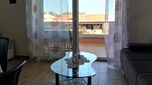 A living room with a glass table, a sofa, and a view of a residential area through a window with patterned curtains. - One bedroom apartment with seaview balcony (Nin)