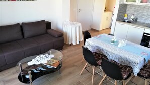 A living room with a dark brown sofa, a glass coffee table, and a dining table with chairs. A small round table with a white tablecloth is in the background. - One bedroom apartment with seaview balcony (Nin)