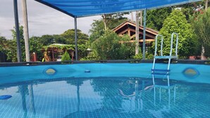 Una piscina azul cristalina con una escalera, rodeada de vegetación exuberante y una estructura de madera al fondo.