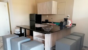 Una cocina compacta con encimera negra, pileta integrada y cafetera. Hay zonas de estar con cojines grises y un mueble de madera sobre la encimera.