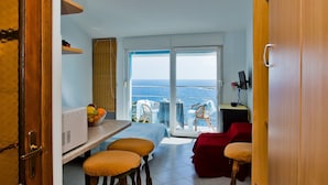 Espaço compacto com cozinha americana, área de refeições com banquetas, sofá vermelho e varanda com vista para o mar.