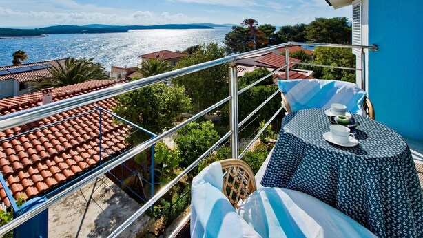 Varanda com mesa posta para dois, com vista para uma área costeira com telhados de terracota e para o mar.
