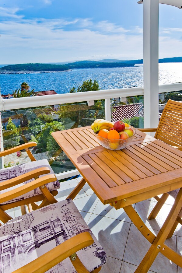 A balcony with a table and chairs, a bowl of fruit, and a view of the sea and greenery.
