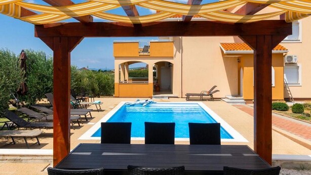 A pool area with a pergola overhead, lounge chairs, and a view of a building with a yellow facade.