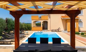 A pool area with a pergola overhead, lounge chairs, and a view of a building with a yellow facade. - Four bedroom villa with private pool (Sukosan)