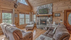 A log cabin interior with a stone fireplace, wooden furniture, and a television mounted on the wall.