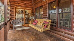 A wooden porch with a swing seat and a wicker chair, a stone fireplace, and large windows.