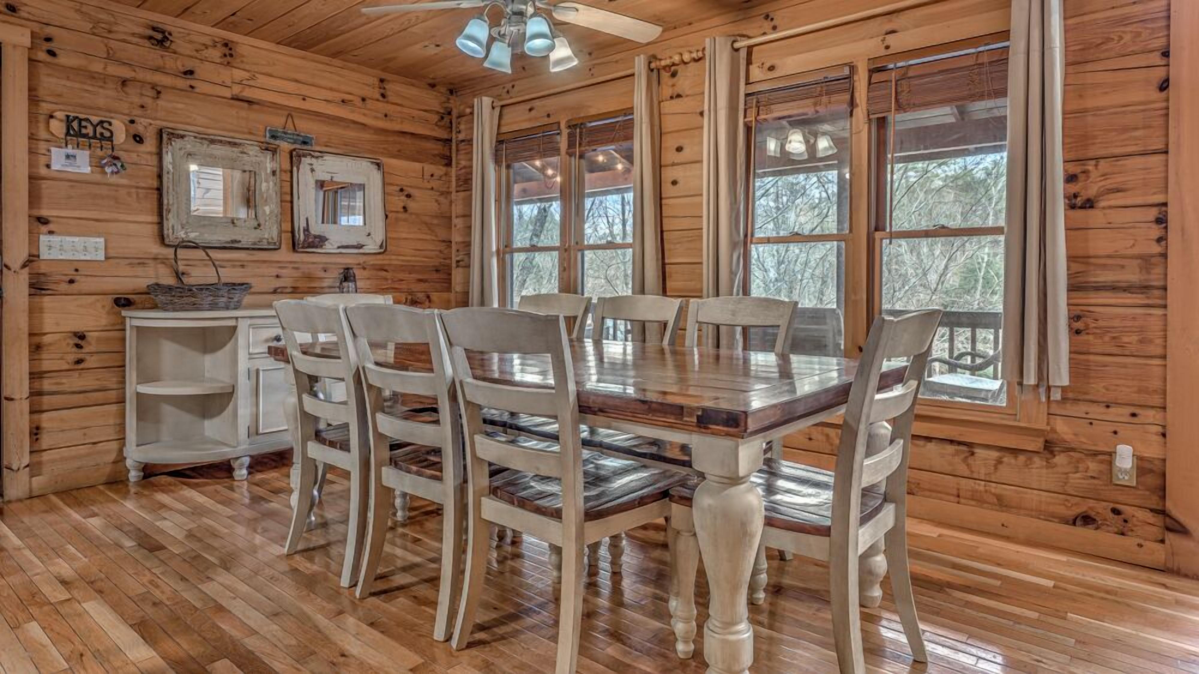 Eine Holzhütte mit Essbereich, der einen Holztisch, Stühle, große Fenster und einen Deckenventilator umfasst.