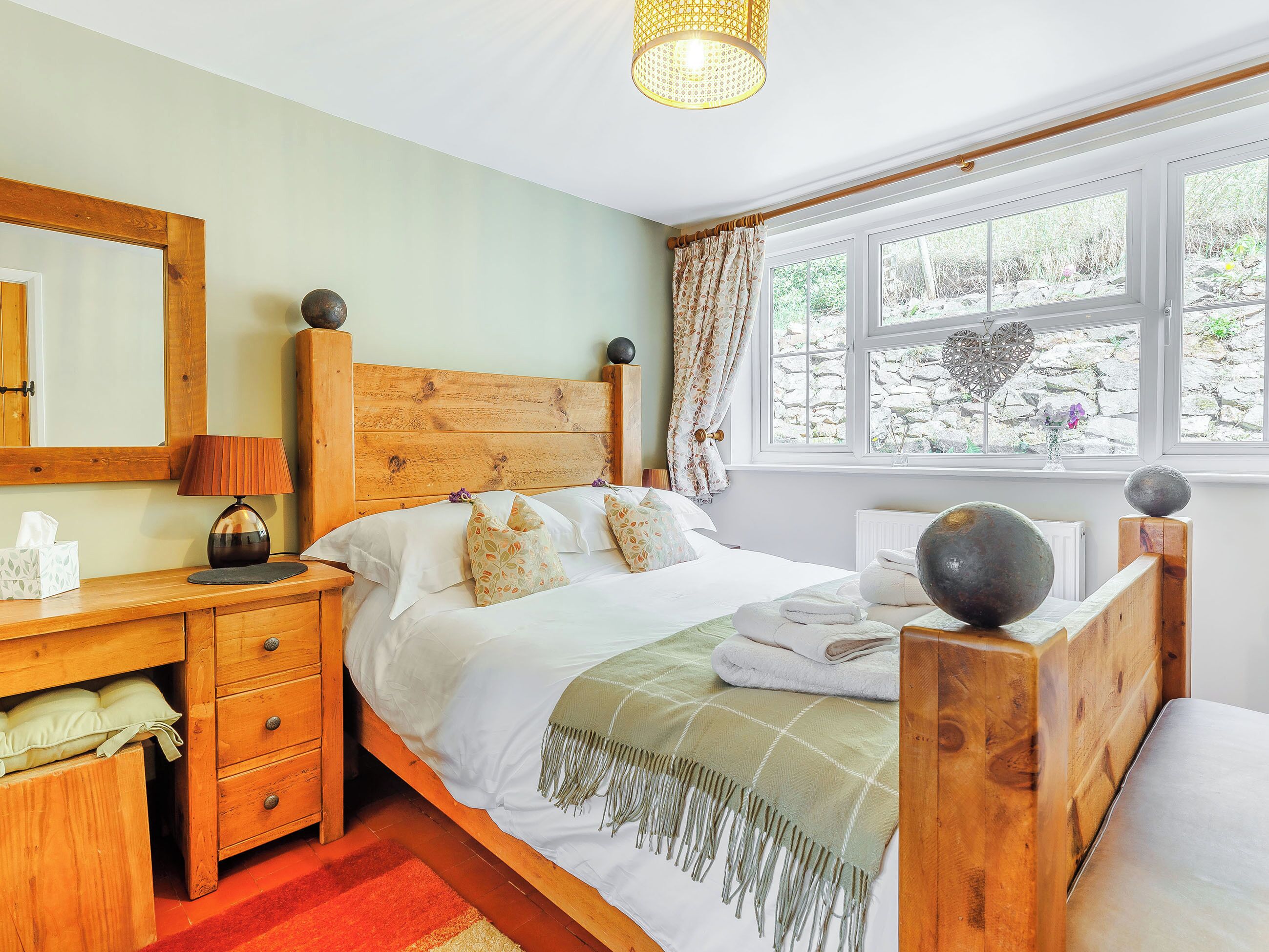 A bedroom with a wooden headboard, a bedside table with a lamp, a window with curtains, and a view of the outdoors.