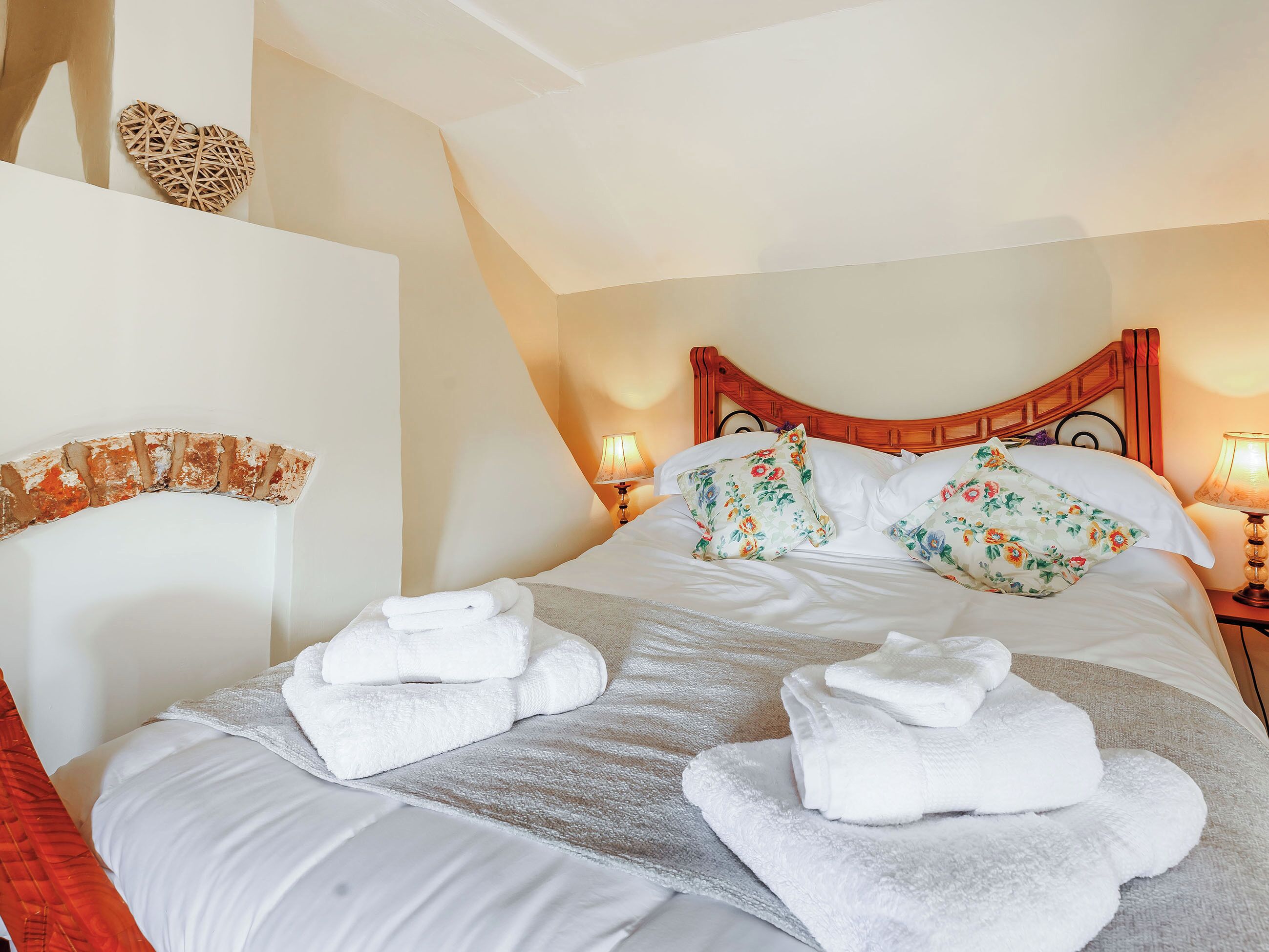 A neatly made bed with floral pillows and white towels, a wooden headboard, and a wall-mounted heart decoration.