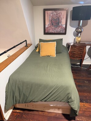 A bedroom with a wooden headboard, a bed with green bedding, a yellow pillow, a wooden nightstand with a lamp, and a framed artwork on the wall. - Darling cottage near entrance to Letchworth State Park  overlooking Silver Lake! (Castile)