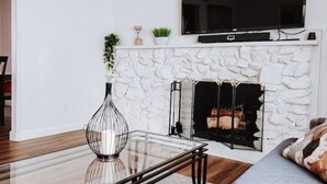 A modern living room with a fireplace, a flat-screen TV mounted above it, and a glass coffee table. - Beautiful and Cozy 4-BR residential home! (Oxnard)