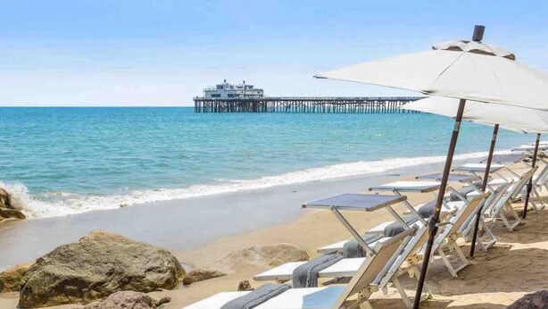 A beach with lounge chairs and umbrellas, a pier in the background, and a building on the pier. - Beautiful and Cozy 4-BR residential home! (Oxnard)