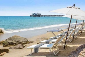 Una playa con sillas y sombrillas, un muelle al fondo y un edificio sobre el muelle.