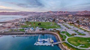 An aerial view of a coastal town with a marina, residential areas, and a hilly landscape in the background. - Beautiful and Cozy 4-BR residential home! (Oxnard)