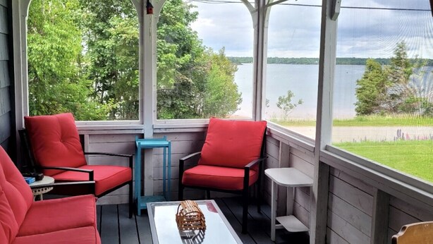 A covered porch with red chairs, a small table, and a view of a lake. - Quaint 2 bedroom water view cottage.  Near great golfing, beaches and breweries. (Lower Montague)