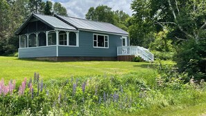 A single-story house with a porch, surrounded by a grassy yard and wildflowers. - Quaint 2 bedroom water view cottage.  Near great golfing, beaches and breweries. (Lower Montague)