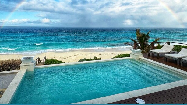 Une vue depuis la piscine donnant sur une plage, avec un arc-en-ciel, une terrasse en bois et des chaises longues.