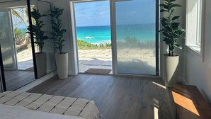 Une chambre moderne dotée d’une grande porte-fenêtre coulissante donnant sur la plage. On y trouve deux plantes en pot et un banc en bois.
