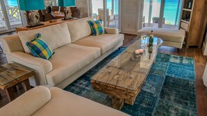 Un salon spacieux avec un canapé beige, une table basse en verre et une vue sur l’océan grâce à de grandes fenêtres.