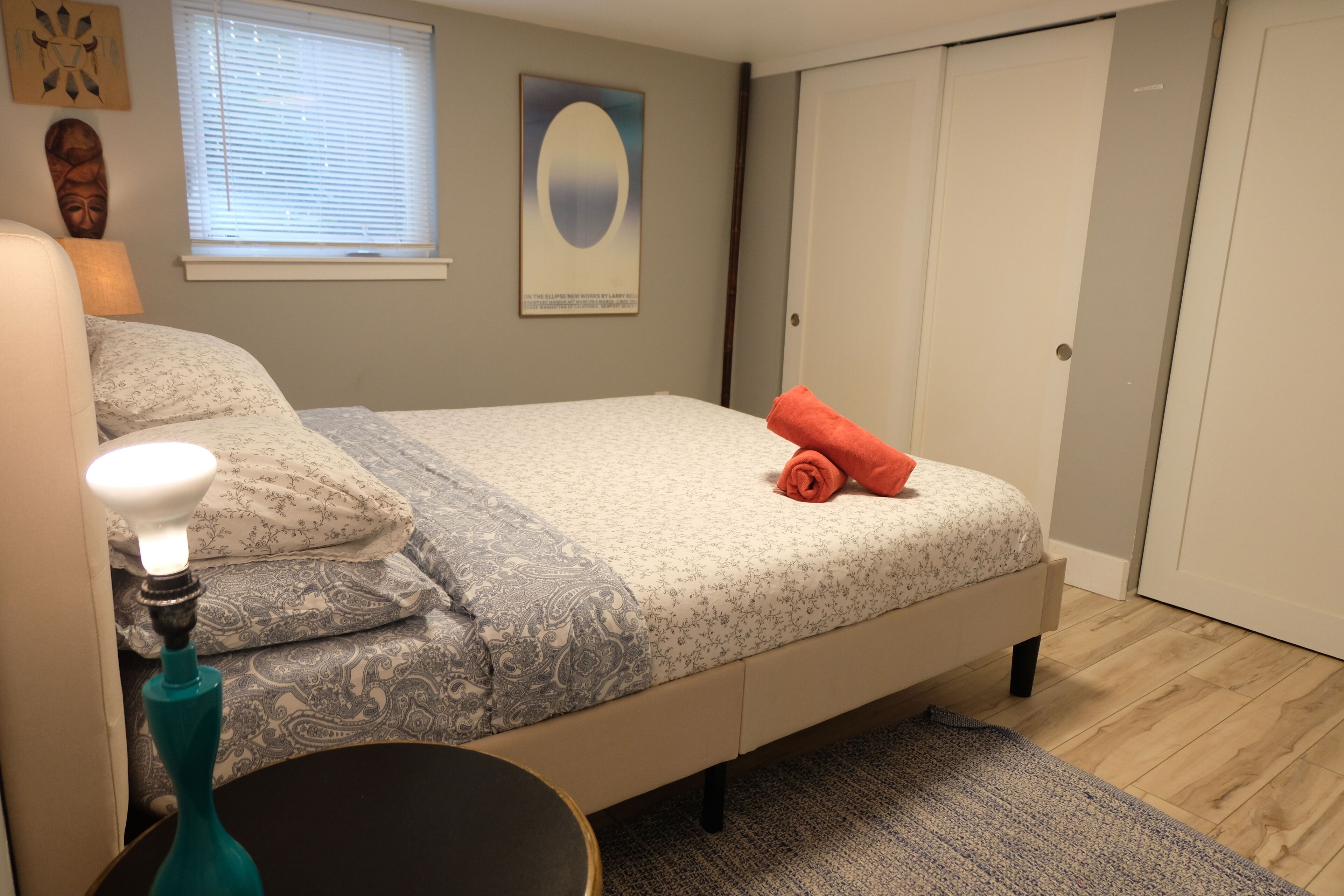 Une chambre à coucher comprenant un lit, une lampe de chevet, une œuvre d’art accrochée au mur, une fenêtre avec des stores et une armoire.