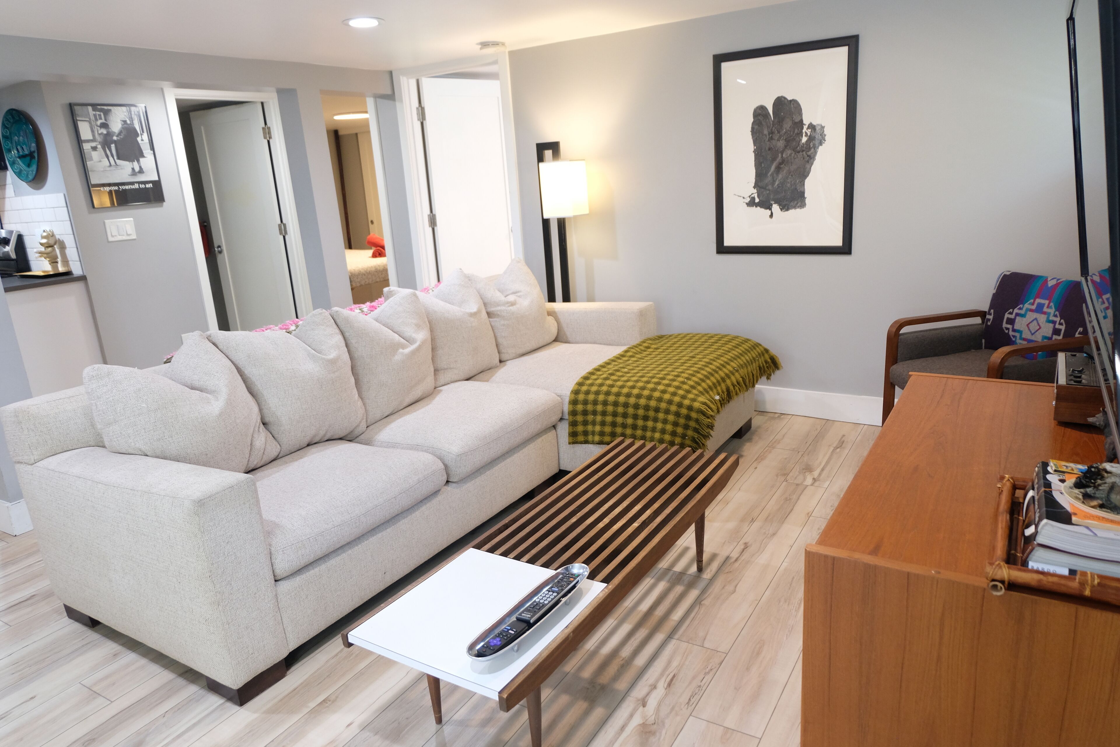 A living room with a sofa, a wooden bench, a coffee table with a remote control, a lamp, and a framed artwork on the wall.